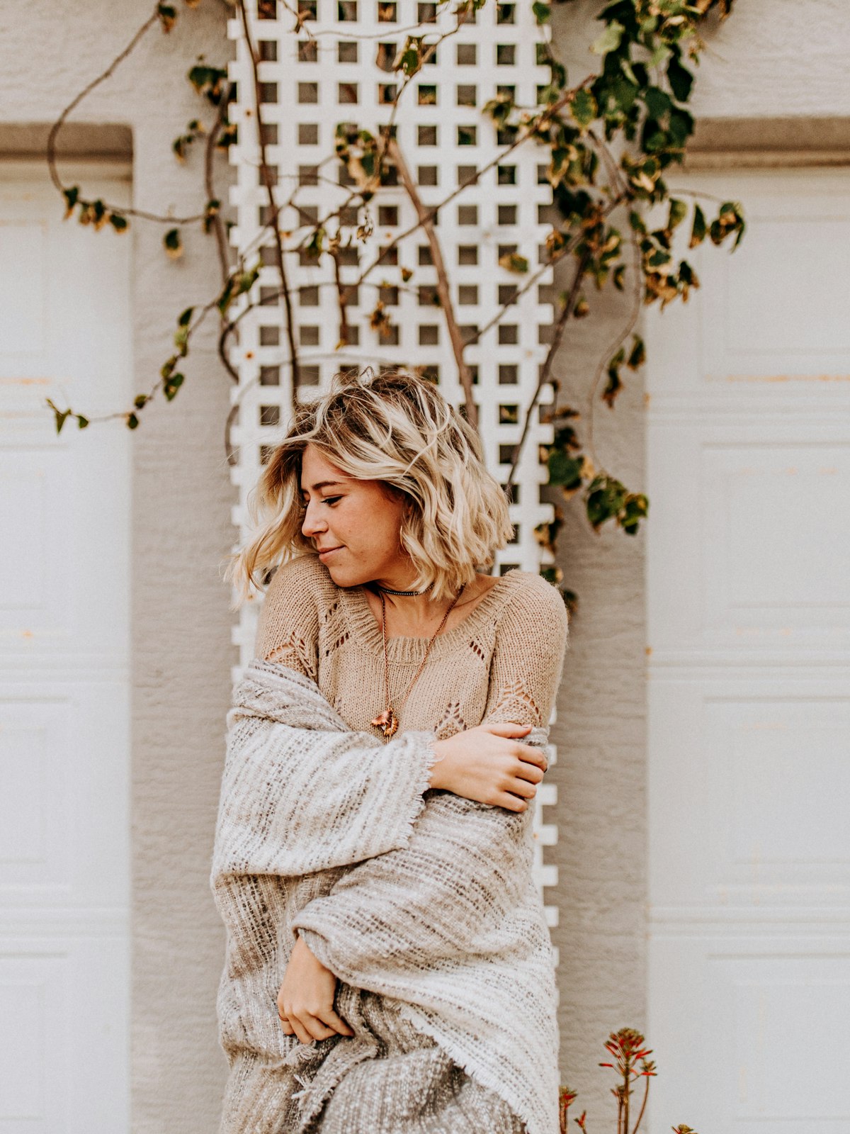 Woman wrapped in a soft oatmeal knit cardigan against a white trellis